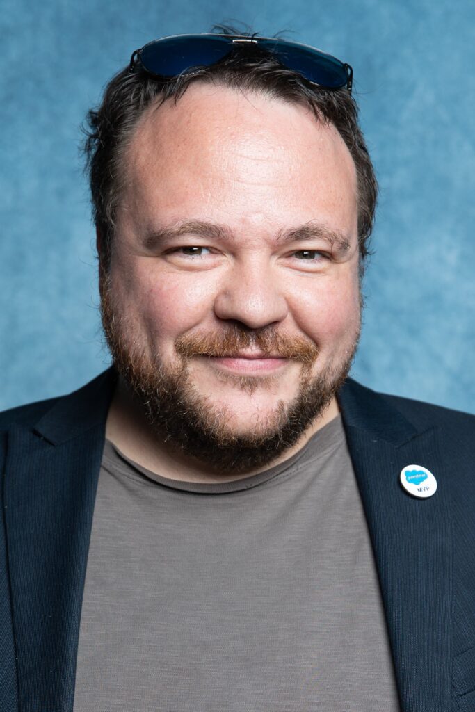 A headshot of a smiling man with short, dark hair and a beard. He’s wearing a dark blazer over a casual shirt and has sunglasses resting on top of his head. The background is a solid blue, and he has a Salesforce pin on his blazer.