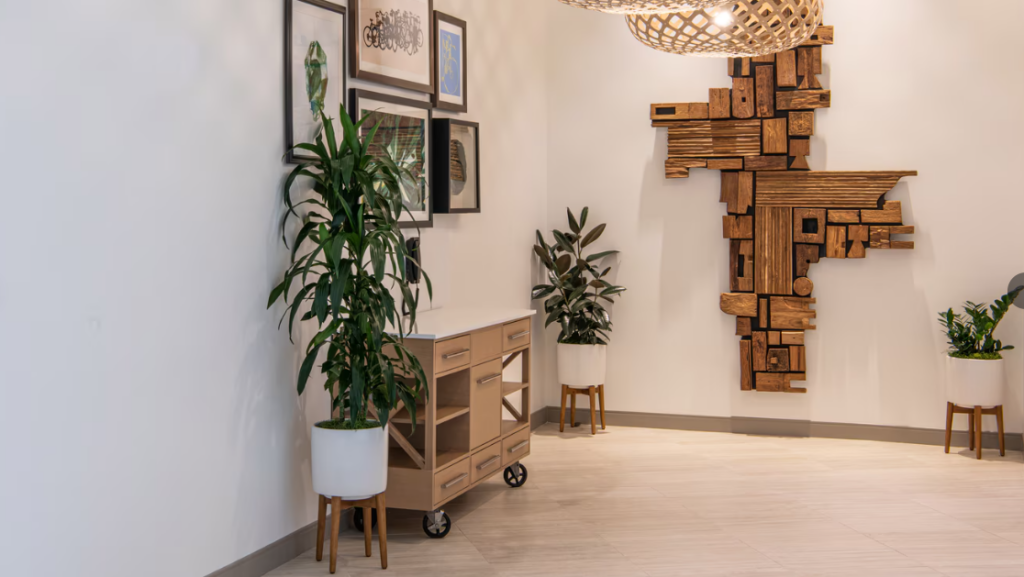 An indoor space with modern decor, featuring potted plants, framed artwork on the wall, and a wooden sculpture with an abstract design mounted on the wall. A small cart with open shelving is positioned against the wall beneath the artwork, and a woven light fixture hangs from the ceiling.