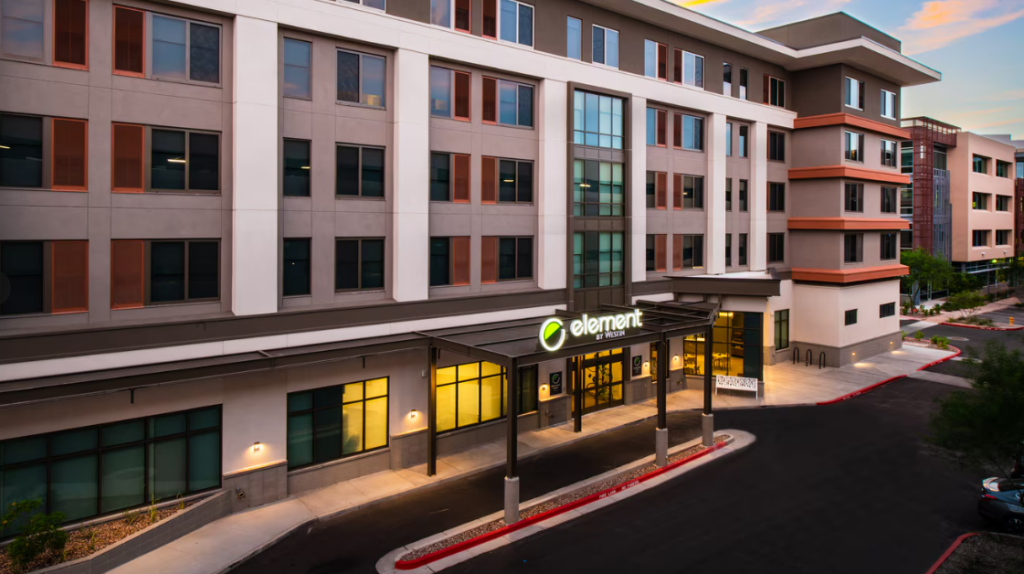 Exterior of the Element by Westin hotel at dusk, with warm lights illuminating the entrance. The modern building features large windows, a covered entryway, and a green Element logo sign above the doors.