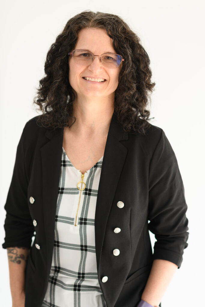 A smiling woman with curly hair and glasses, wearing a black blazer with silver buttons over a checkered top, standing against a white background.