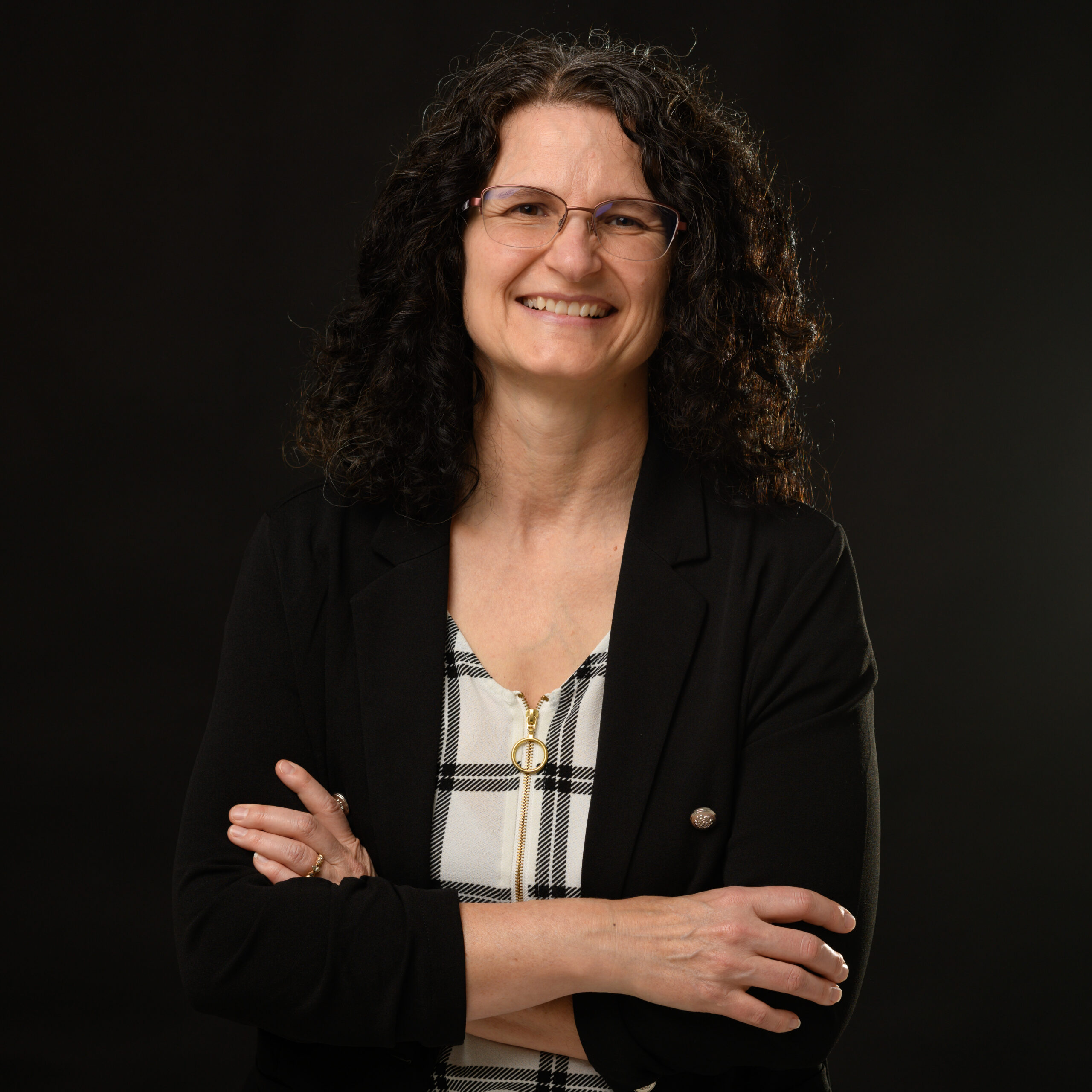 A professional woman with curly hair and glasses, wearing a black blazer over a checkered top, smiling confidently with her arms crossed against a dark background.