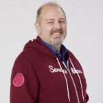 A smiling man with a beard wearing a maroon hoodie that reads 'Service Champion' and a blue shirt underneath, standing with hands in his pockets against a light background.