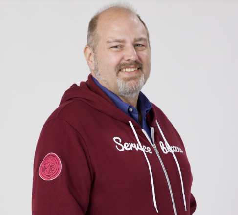 A smiling man with a beard wearing a maroon hoodie that reads 'Service Champion' and a blue shirt underneath, standing with hands in his pockets against a light background.