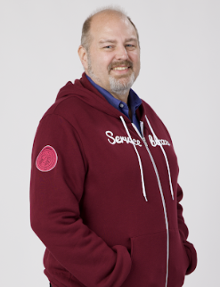 A smiling man with a beard wearing a maroon hoodie that reads 'Service Champion' and a blue shirt underneath, standing with hands in his pockets against a light background.