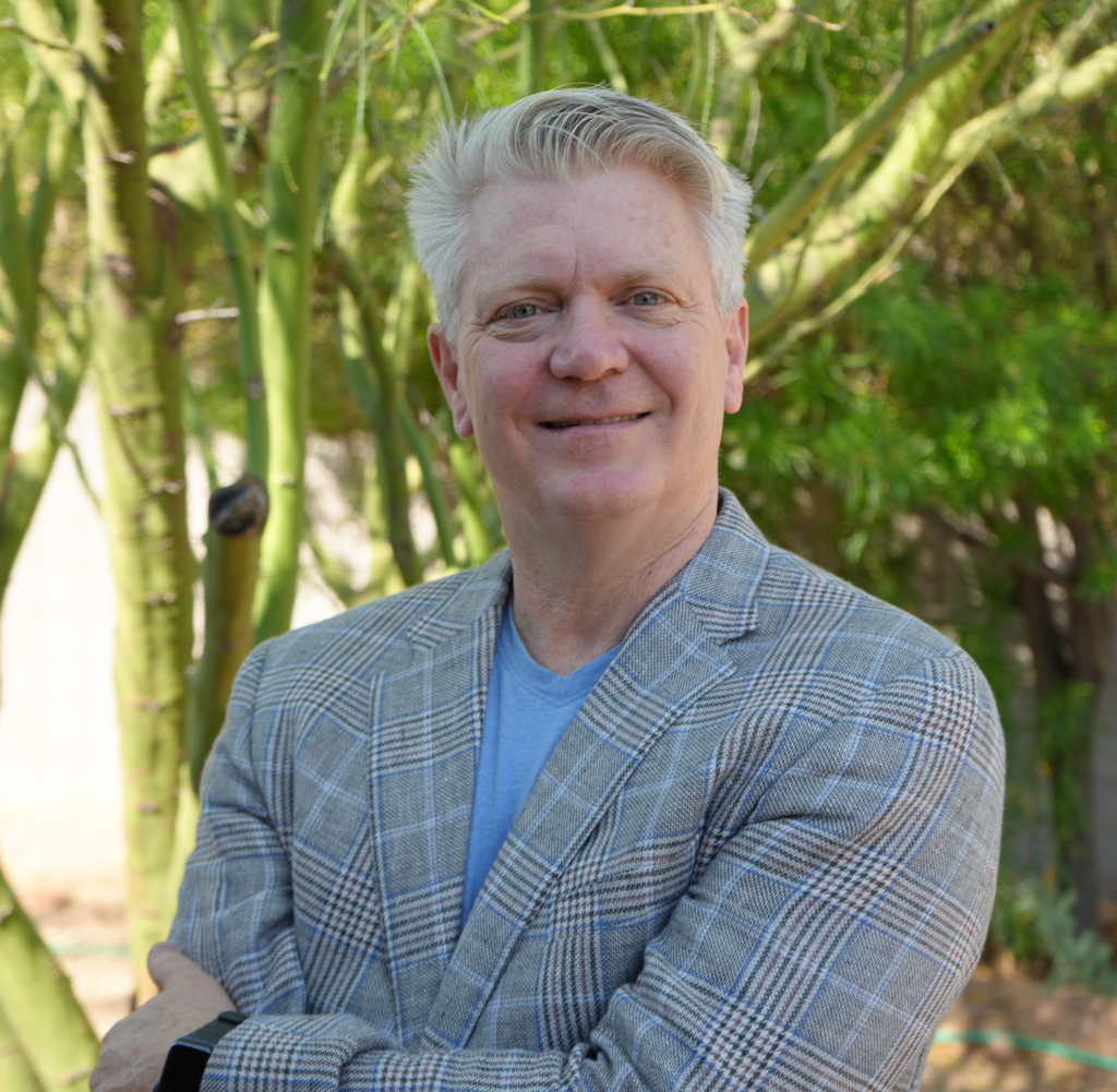 Smiling man in a light plaid blazer and blue shirt, standing outdoors with green trees and sunlight in the background.