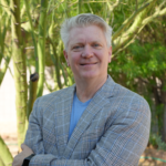 Smiling man in a light plaid blazer and blue shirt, standing outdoors with green trees and sunlight in the background.