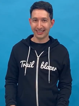 A man smiling and wearing a black hoodie with the words 'Trail Blazer' on the front, standing against a bright blue background.