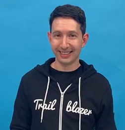 A man smiling and wearing a black hoodie with the words 'Trail Blazer' on the front, standing against a bright blue background.
