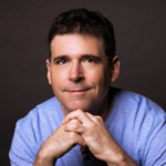 A man with short dark hair wearing a light blue shirt, looking directly at the camera with a slight smile, hands clasped under his chin, against a dark background