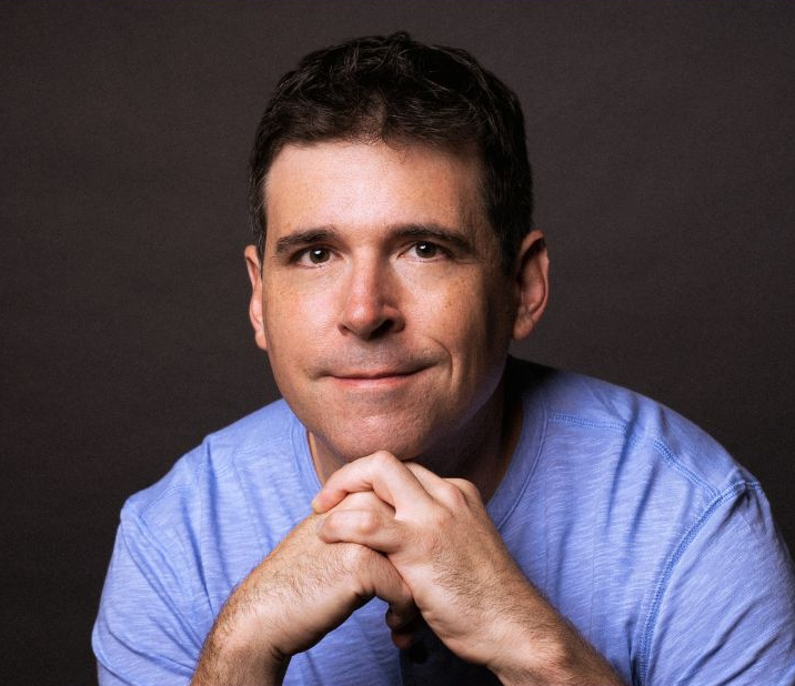 A man with short dark hair wearing a light blue shirt, looking directly at the camera with a slight smile, hands clasped under his chin, against a dark background